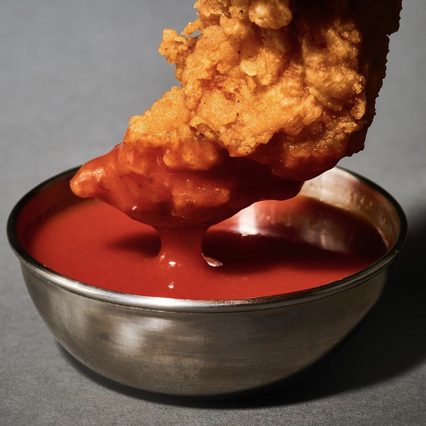 Chicken tender being dipped into a bowl of Rickey's hot sauce on a gray background