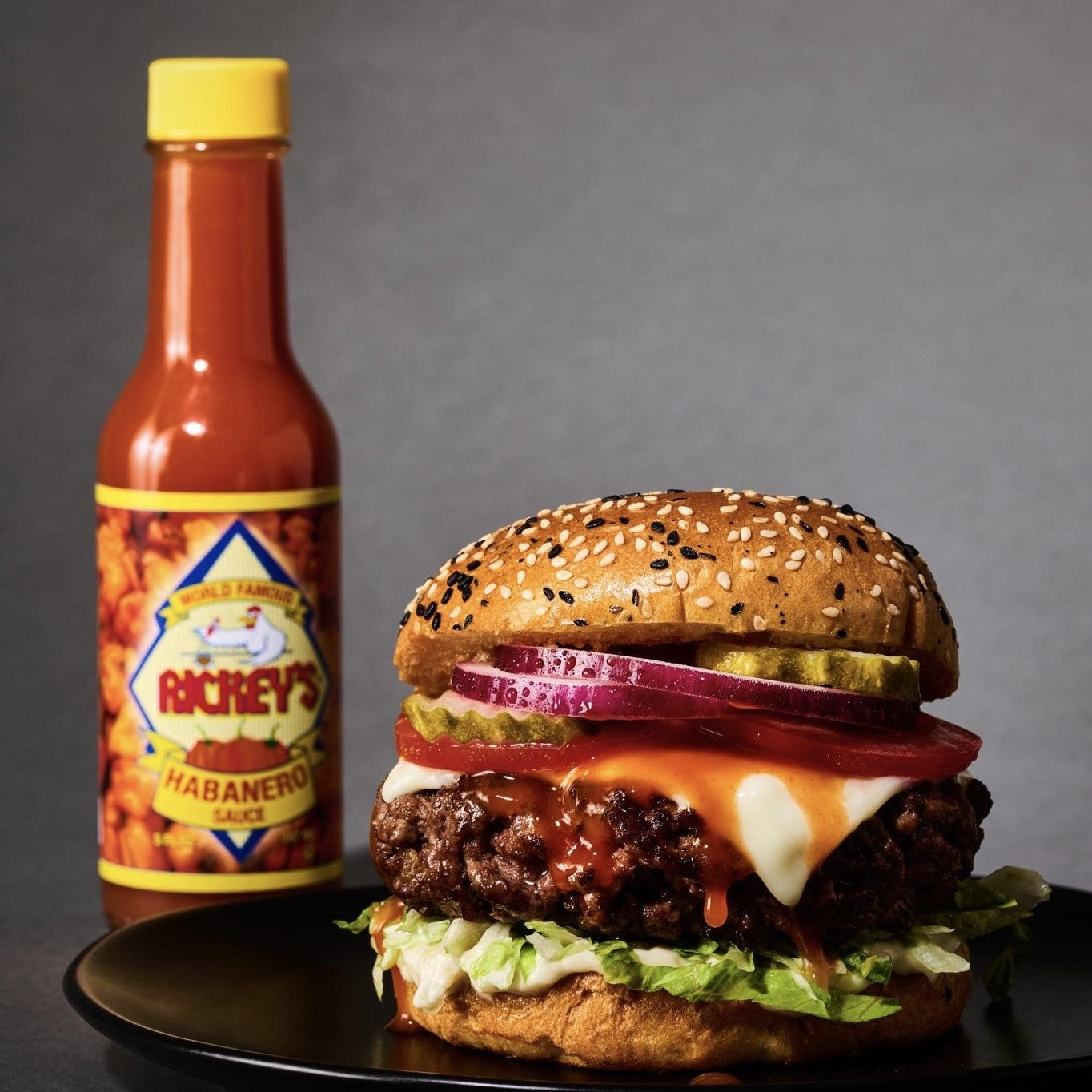 Hamburger with cheese, lettuce, and red onion on a sesame seed bun next to a bottle of Rickey's Habanero sauce.